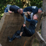 Two men pulling a third man over a tilted wall